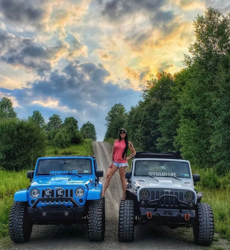 Two Jeeps with offroad life stickers on a dirt road with a girl standing between them, surrounded by trees and a cloudy sky.