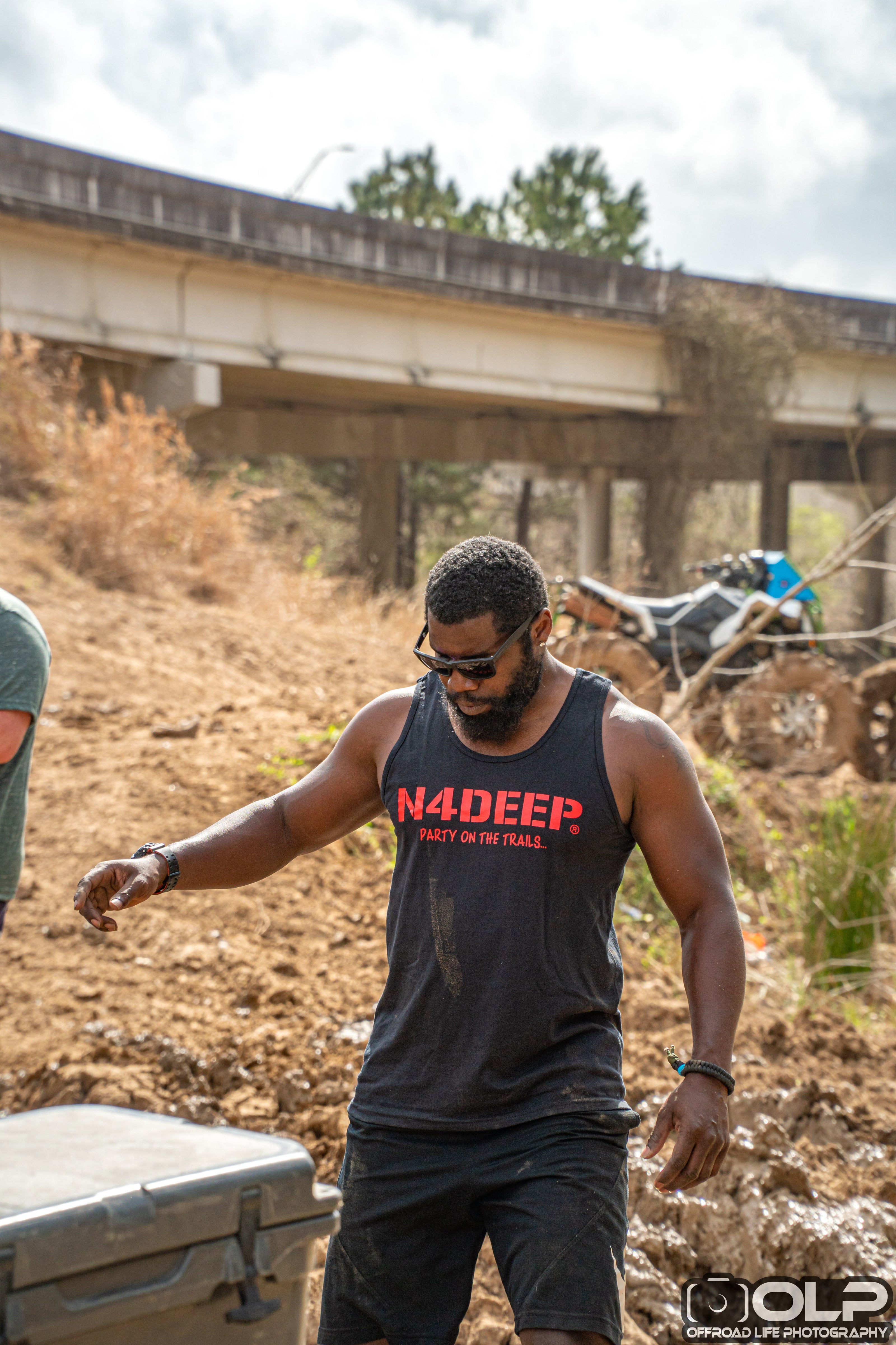 Man wearing a 'N4DEEP' tank top in an outdoor setting with a atv and overpass in the background.